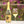 A bottle of La Mesa Aria sparkling white wine with a gold foil and yellow label stands next to a clear wine glass filled with pale golden wine, set on a terrasse table with a blurred garden background of green foliage and pink flowers.