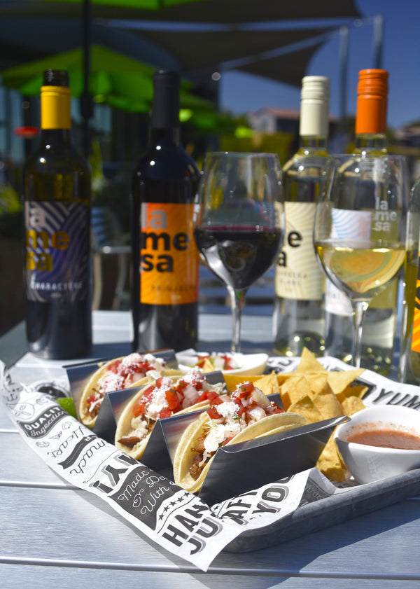 A tray of tacos and tortilla chips on a table outdoors, with wine bottles and filled wine glasses in the background. The food is served on branded paper liner, and the bottles behind it show the La Mesa brand.