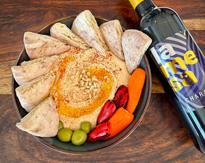 Hummus with pita bread, olives, and vegetables on a wooden table with a bottle of La Mesa Charactere red wine.